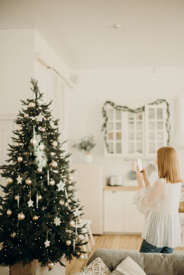 Image of a woman taking a photo of a christmas tree to share on social media. This can be a security breach at christmas time, as burglars can know where expensive gifts are kept under the tree.